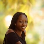 Portrait of a cheerful young woman with braided hair wearing a black top, captured in an outdoor setting with a blurred background.